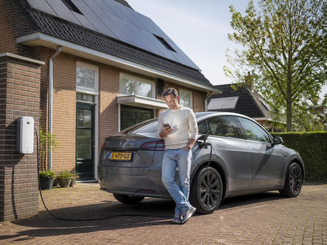 Man met telefoon naast zijn ladende elektrische auto bij een Nederlands huis met zonnepanelen op het dak